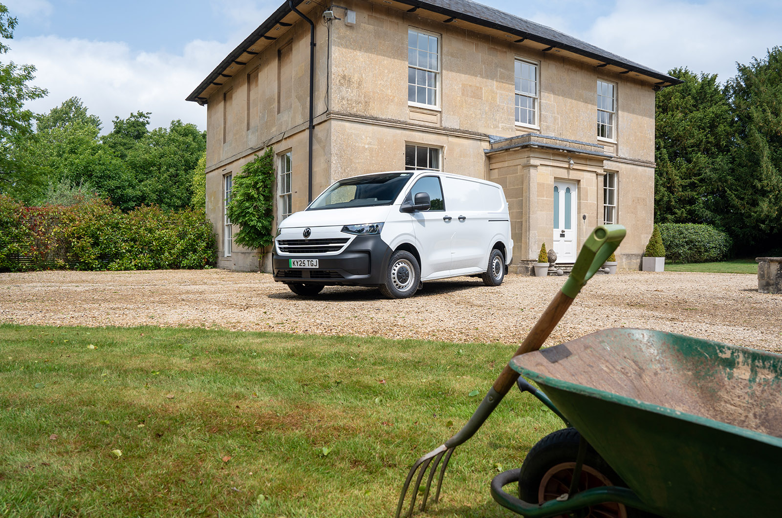 A white Volkswagen Transporter with black bumpers parked outside a country house with a wheelbarrow in the foreground