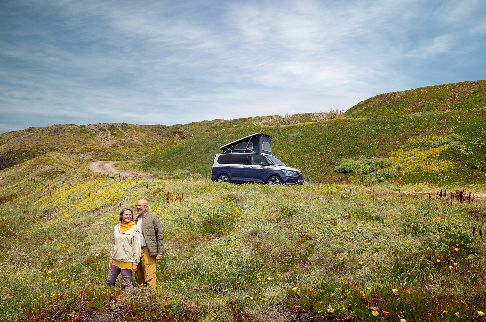 A two-tone blue and silver Volkswagen California Ocean parked up with the roof raised as two passengers admire the view