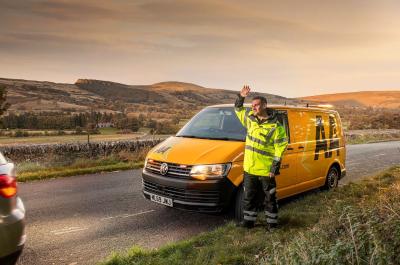 An AA patrol person stands in front of a yellow AA van and waves to his breakdown customers as they drive away