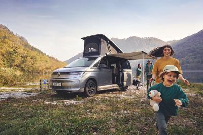 The front left quarter of the Volkswagen California with the pop-up roof and side-awning extended and a child running in the foreground