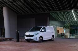 A white Volkswagen Transporter charging while parked outside of a glass-fronted building